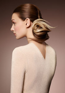 A young woman wearing an ivory, knit, v-neck shirt wears her brown-blonde balayage hair up in a twist ending in a stylized bun.