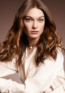 A young woman wearing a ivory blouse showcases her lightly waved, brown hair.