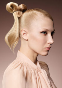 A blonde woman wearing a light pink blouse showcases an knotted up-do.