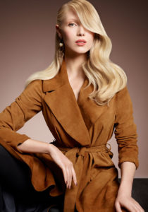 A women wearing a brown trenchcoat showcasing long waved blonde hair swept behind one ear.